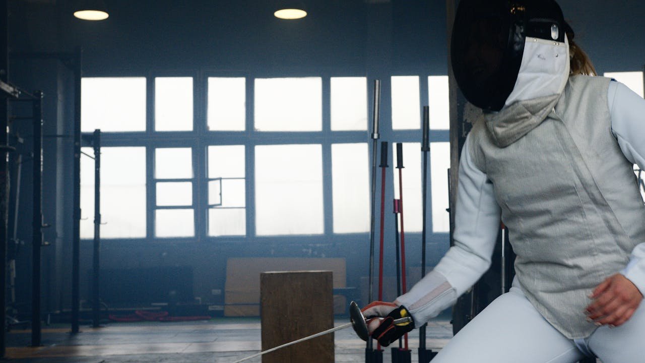 A fencer in protective gear practicing in an indoor salle with fencing swords.
